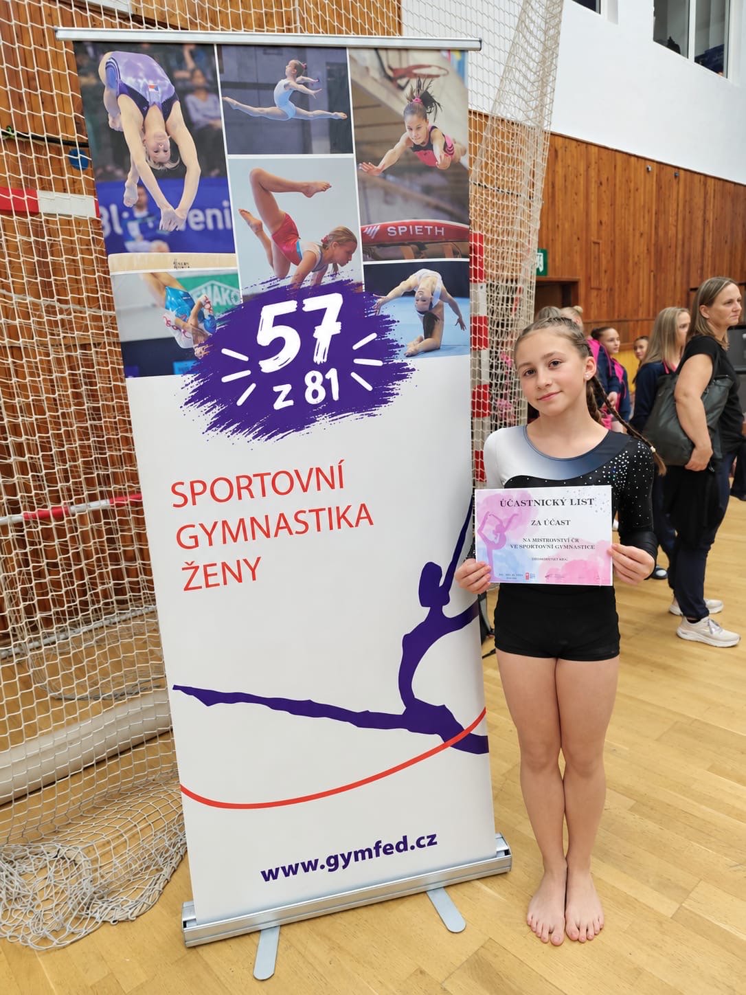 Mladá gymnastka v dresu drží účastnický list vedle reklamního banneru České gymnastické federace v tělocvičně.