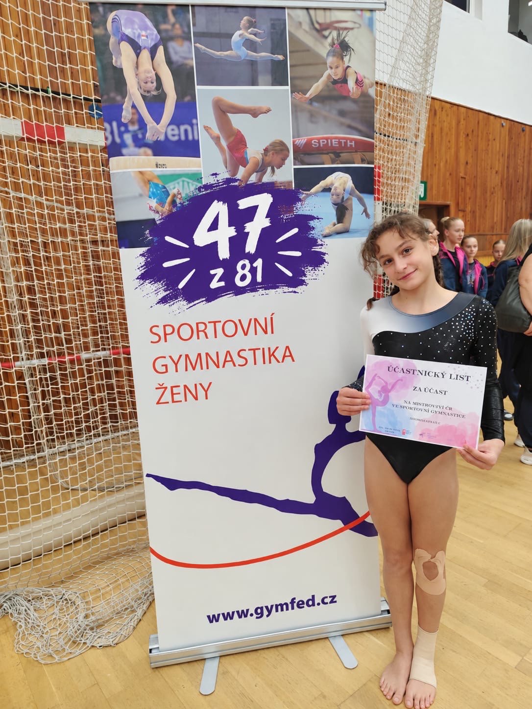Mladá gymnastka v černém dresu drží účastnický list vedle informačního banneru v tělocvičně.