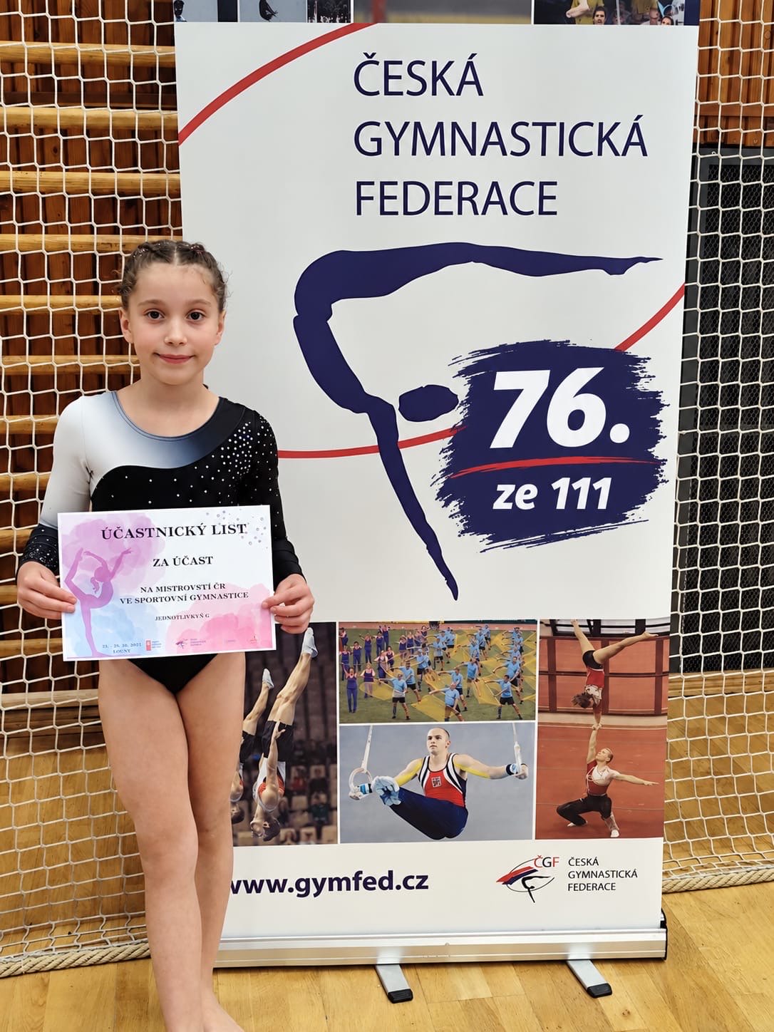 Mladá gymnastka v dresu drží účastnický list vedle banneru České gymnastické federace v tělocvičně.